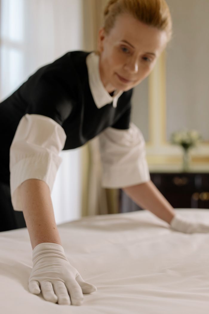 Une gouvernante dispose avec élégance les draps sur un lit dans une chambre d’hôtel luxueuse, illustrant un savoir-faire en matière d’hospitalité.